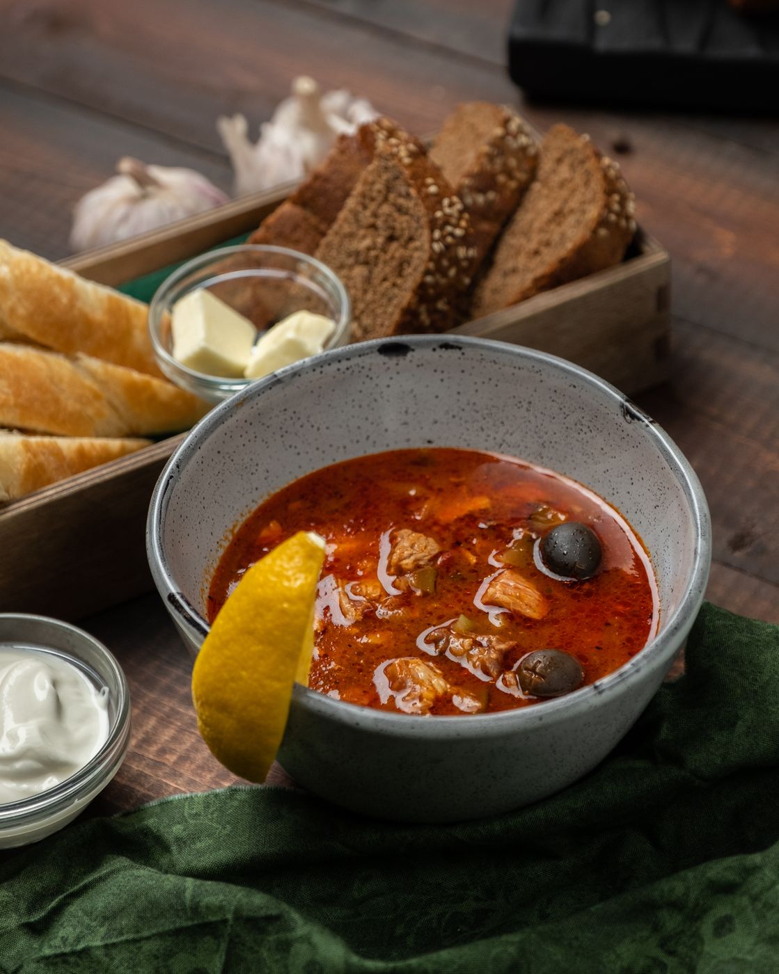 Солянка мясная в серой миске с ломтиком лимона, сметаной и чёрным хлебом