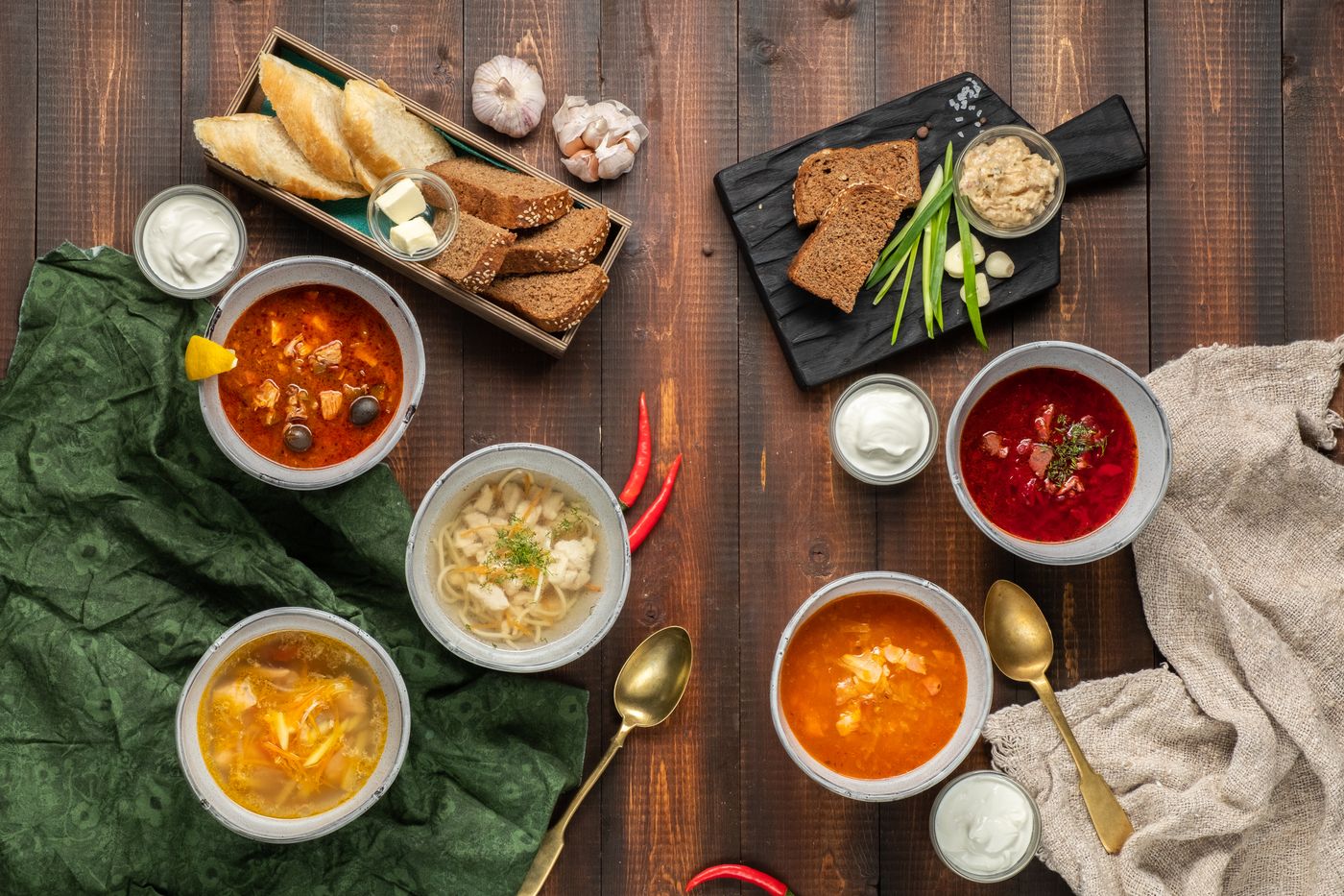 Пять супов на деревянном столе: солянка, борщ, куриный бульон с лапшой, щи — с хлебной корзиной и сметаной